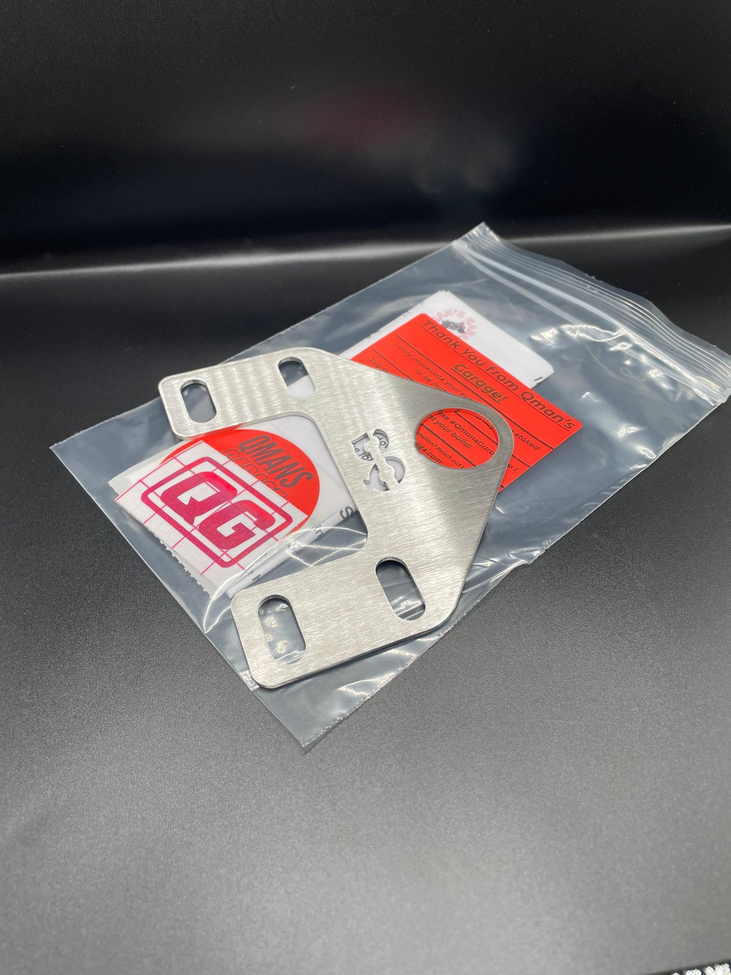 Metal bracket in clear packaging with a red label on a dark surface